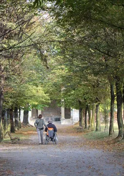 Ein alter Mann sitzt im Rollstuhl und eine Frau geht in einem Park vorbei Ostewind Betreuungs- und Entlastungsdienst